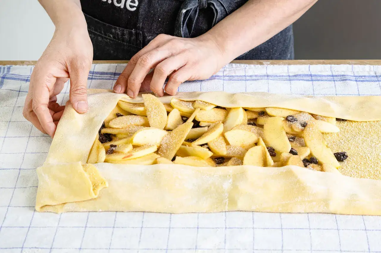 Hände falten den Teig für ein Apfelstrudel Rezept. Äpfel und Rosinen sind bereit zum Backen.