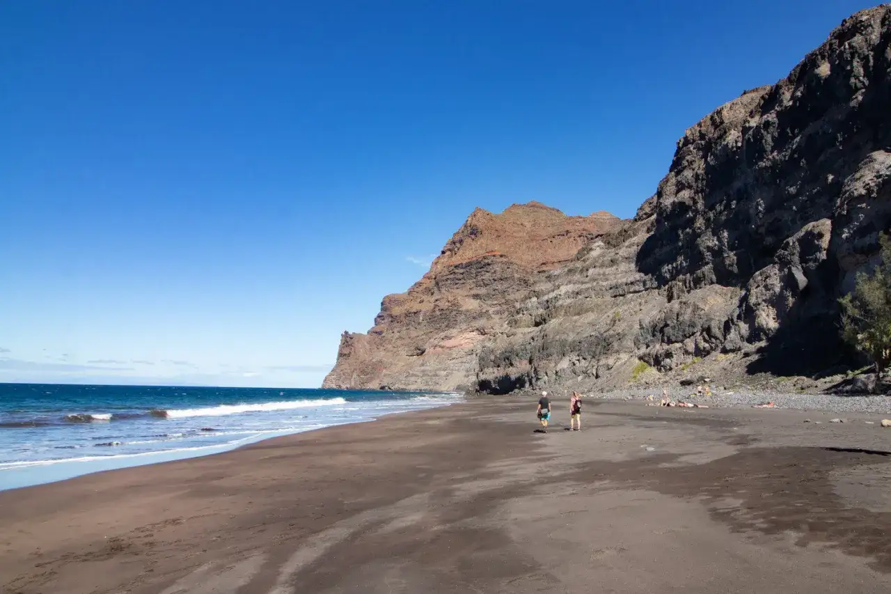 Zwei Personen spazieren am dunklen Sandstrand von Gran Canaria entlang, während im Hintergrund eine imposante Felswand aufragt.