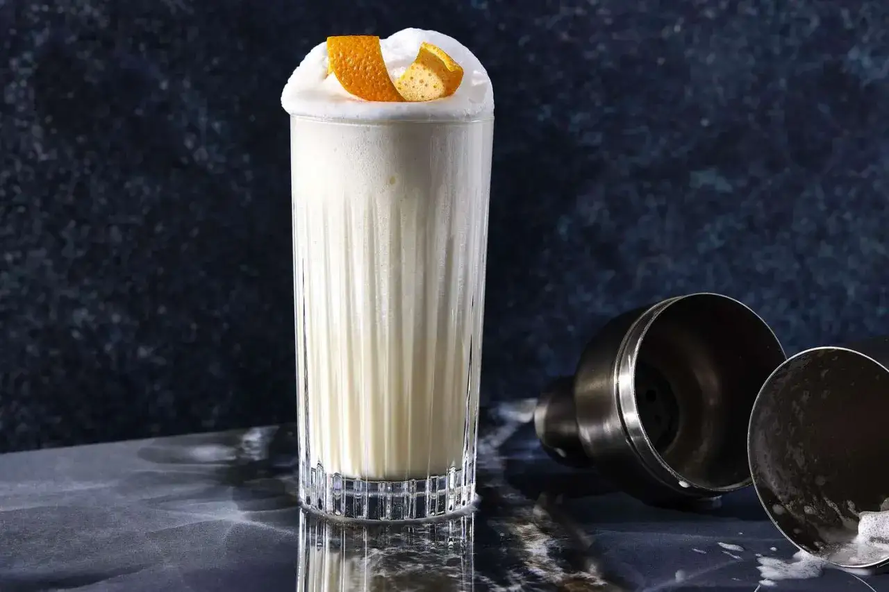 En kr&auml;mig Ramos Gin Fizz med apelsinskal som garnering. Bredvid st&aring;r en cocktailshaker.