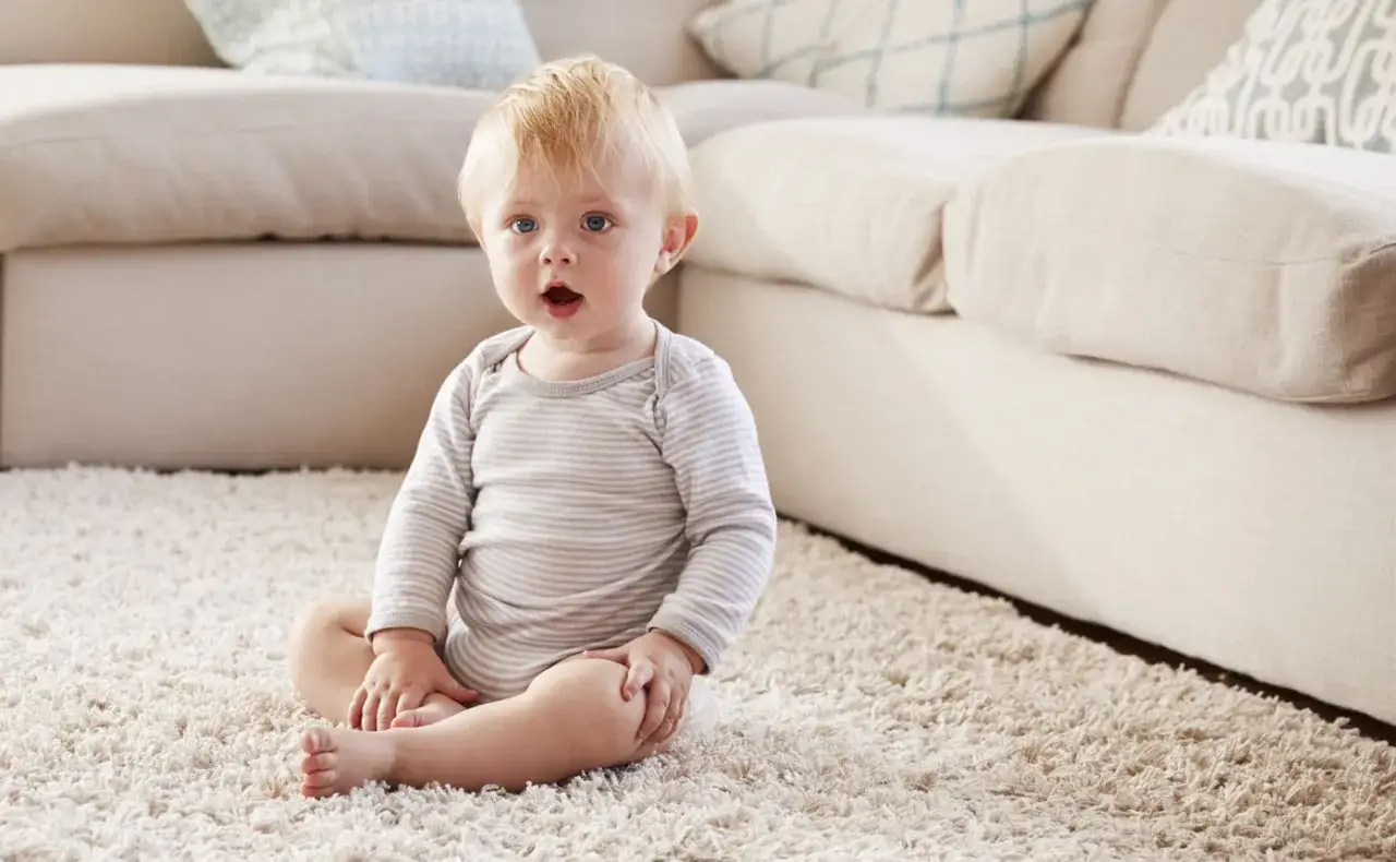 Ein blondes Baby sitzt auf einem flauschigen Teppich vor einem Sofa.