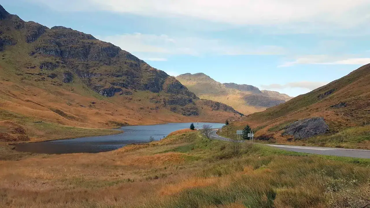Herbstliche Landschaft in Schottland im September. Ein See liegt eingebettet zwischen sanften, braun-grünen Hügeln, eine Straße schlängelt sich vorbei.