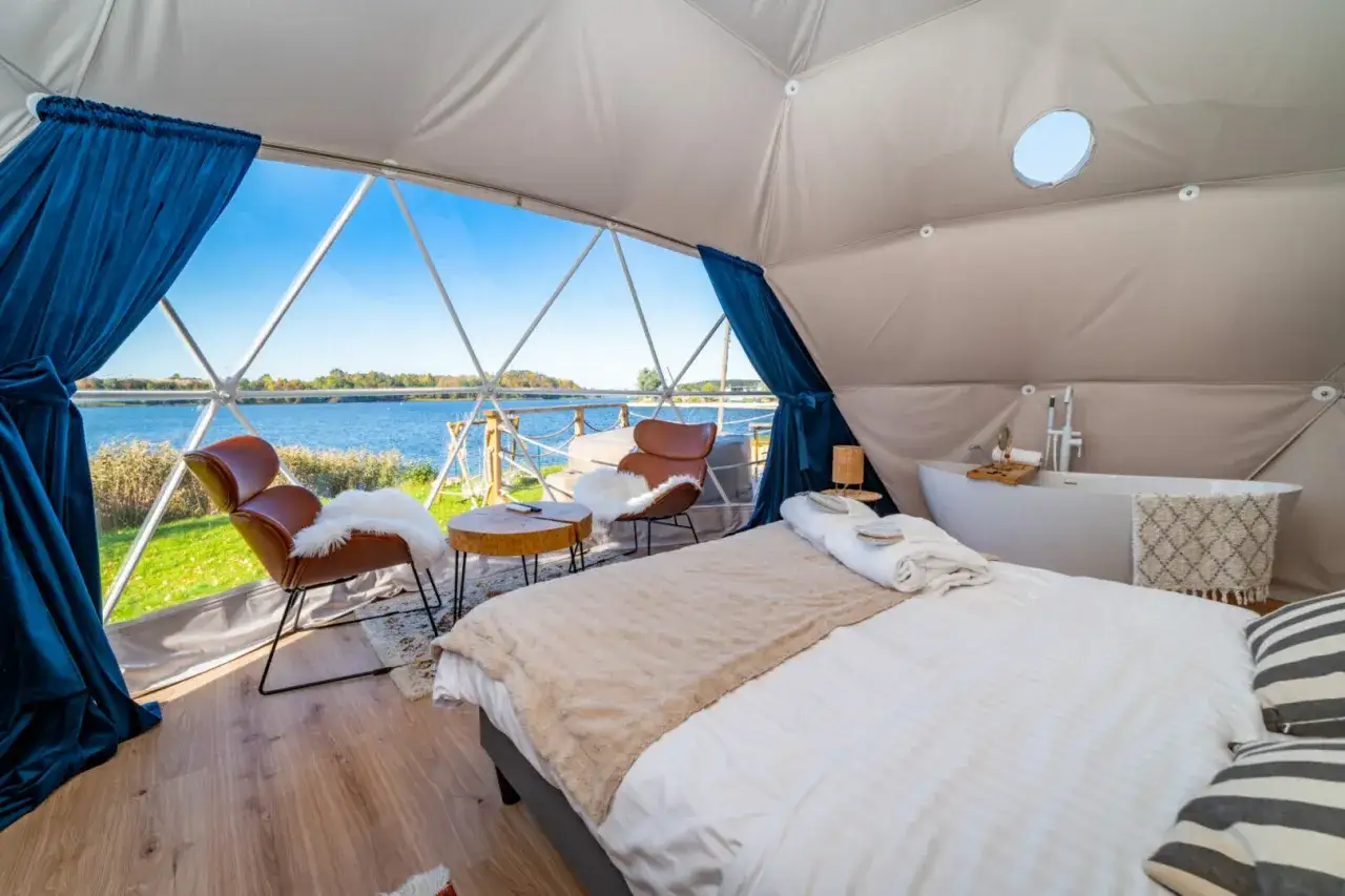 Luksusowy glamping nad jeziorem: wygodne łóżko, wanna i fotele z widokiem na wodę.
