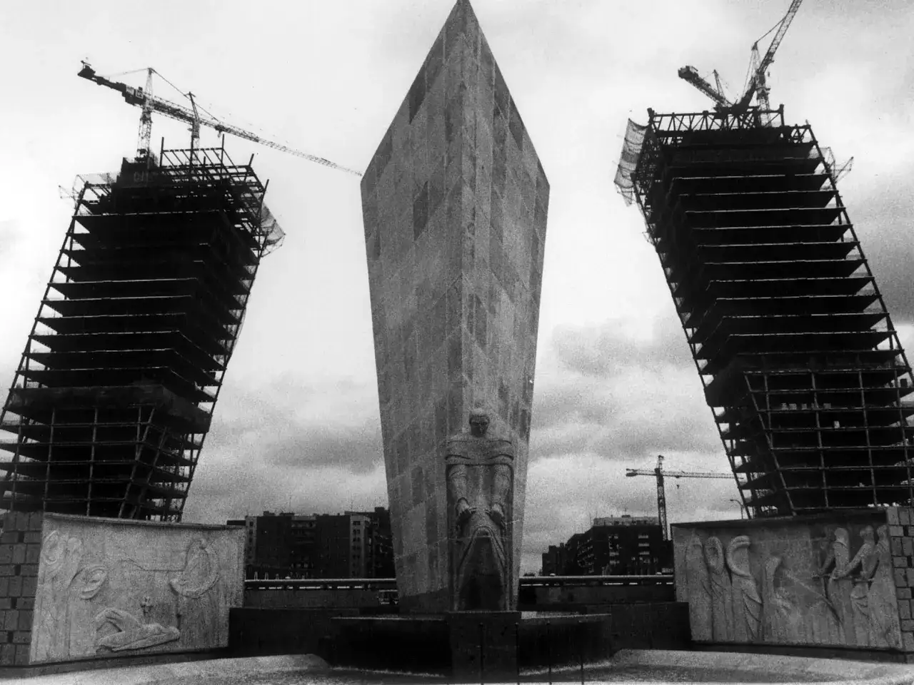 Las **torres KIO** en **construcción**, con grúas y estructuras metálicas visibles, flanquean un obelisco central con una escultura y relieves.