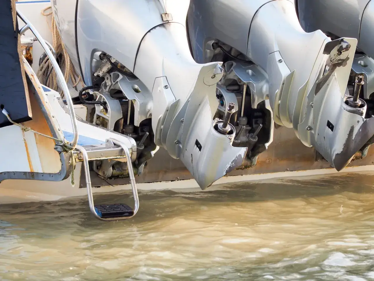 Vue rapprochée des moteurs hors-bord d'un bateau, l'un d'eux semble avoir un problème moteur. L'eau trouble reflète le ciel.