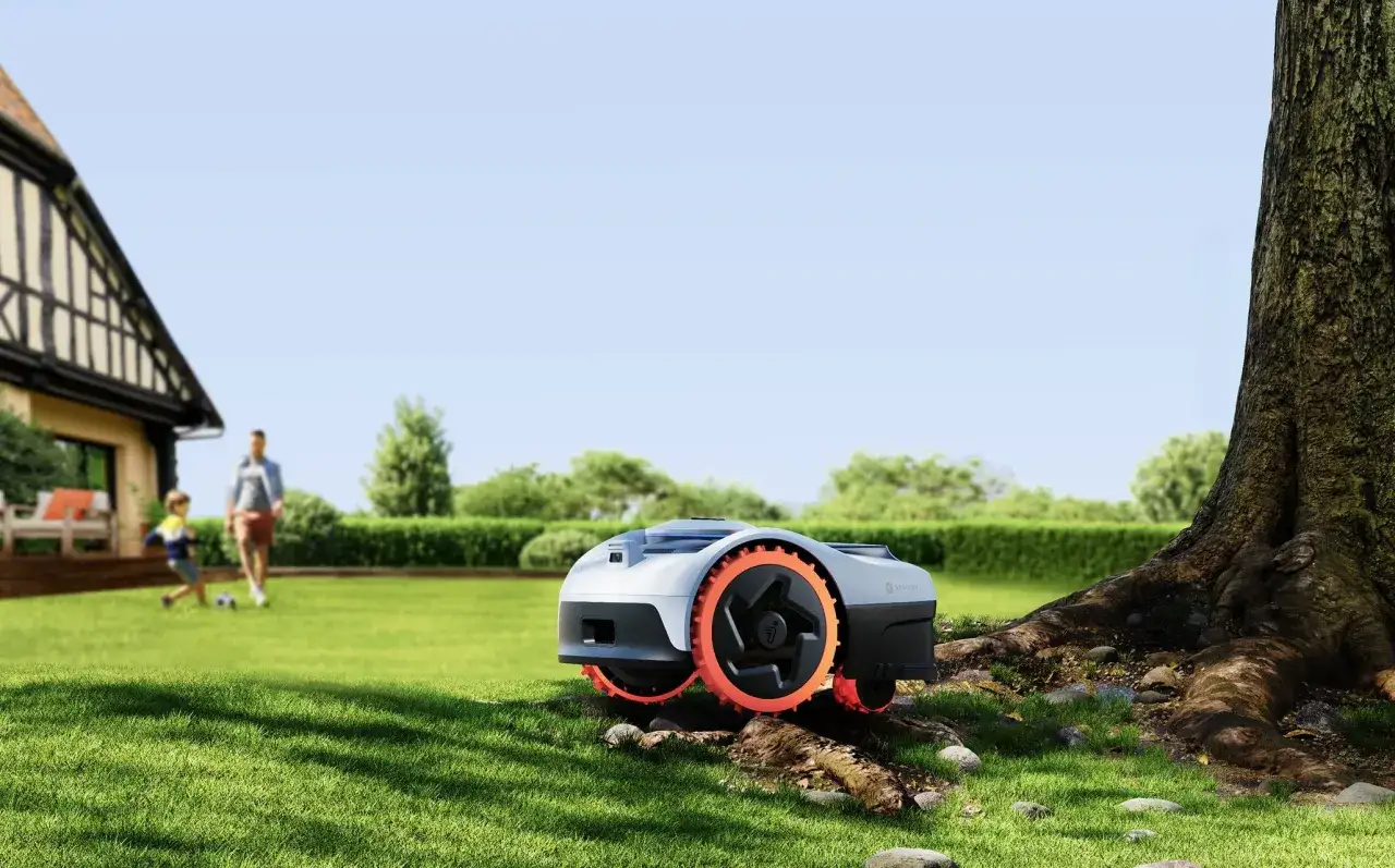 Segway Navimow test i en solig trädgård. En robotgräsklippare med orange hjul står på gräsmattan nära ett stort träd. I bakgrunden leker en pappa och son fotboll.