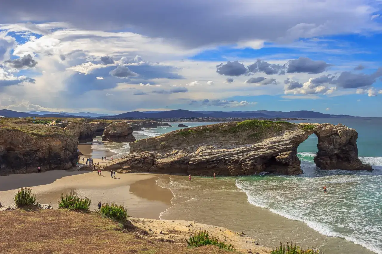 Arco natural en las playas de Galicia, con gente paseando y el mar turquesa bajo un cielo nublado.