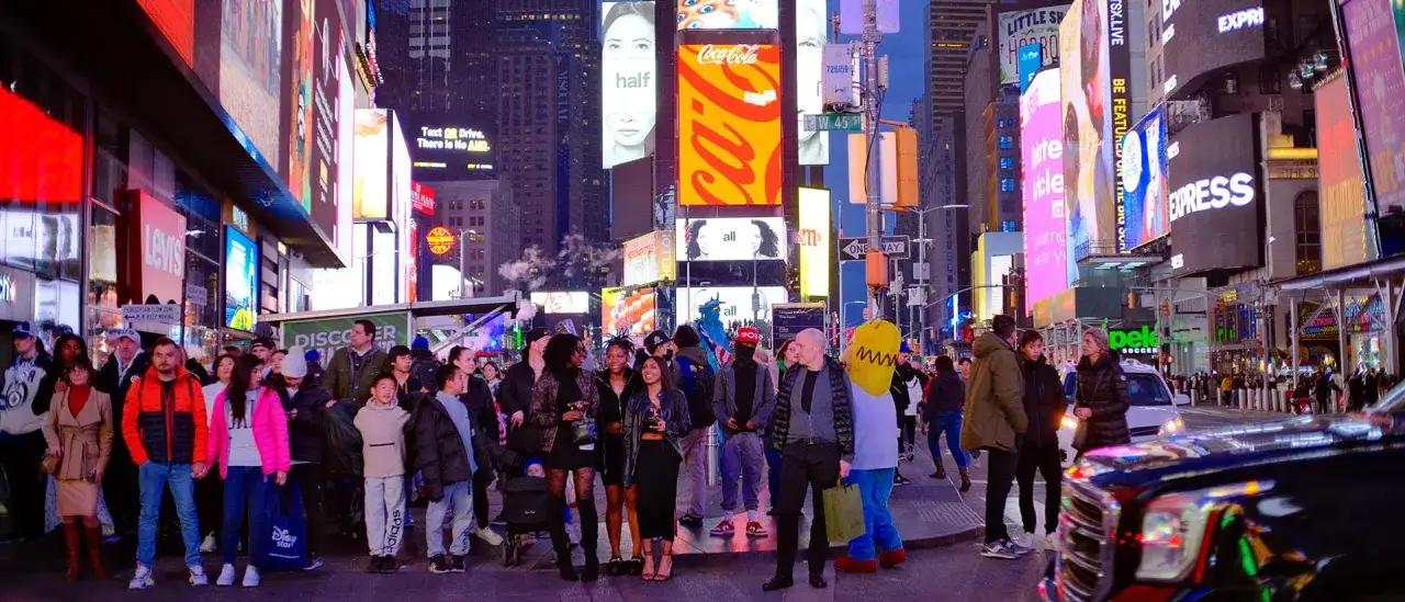 Tętniąca życiem usa street pełna neonów i ludzi. W tłumie widać przebrane postacie, a nad głowami migoczą gigantyczne ekrany reklamowe.
