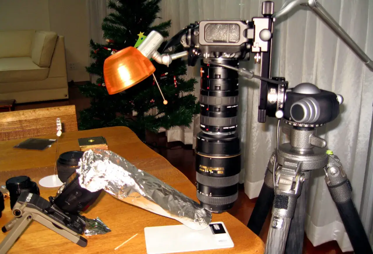 Zestaw do makrofotografii z lampą, obiektywami i statywem na stole, w tle choinka i kanapa.