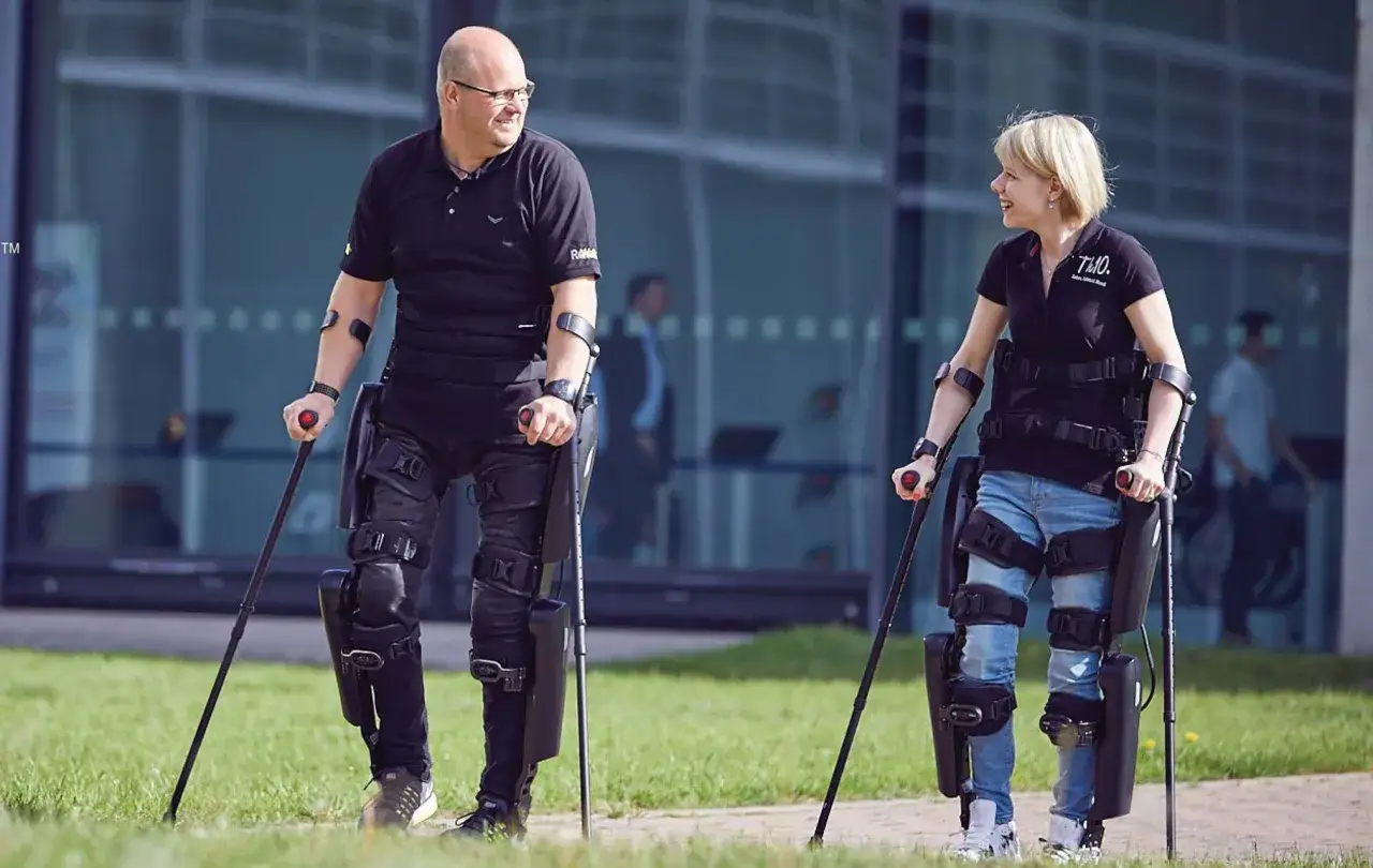 Uomo e donna camminano con un esoscheletro, un supporto robotico che aiuta a muoversi.