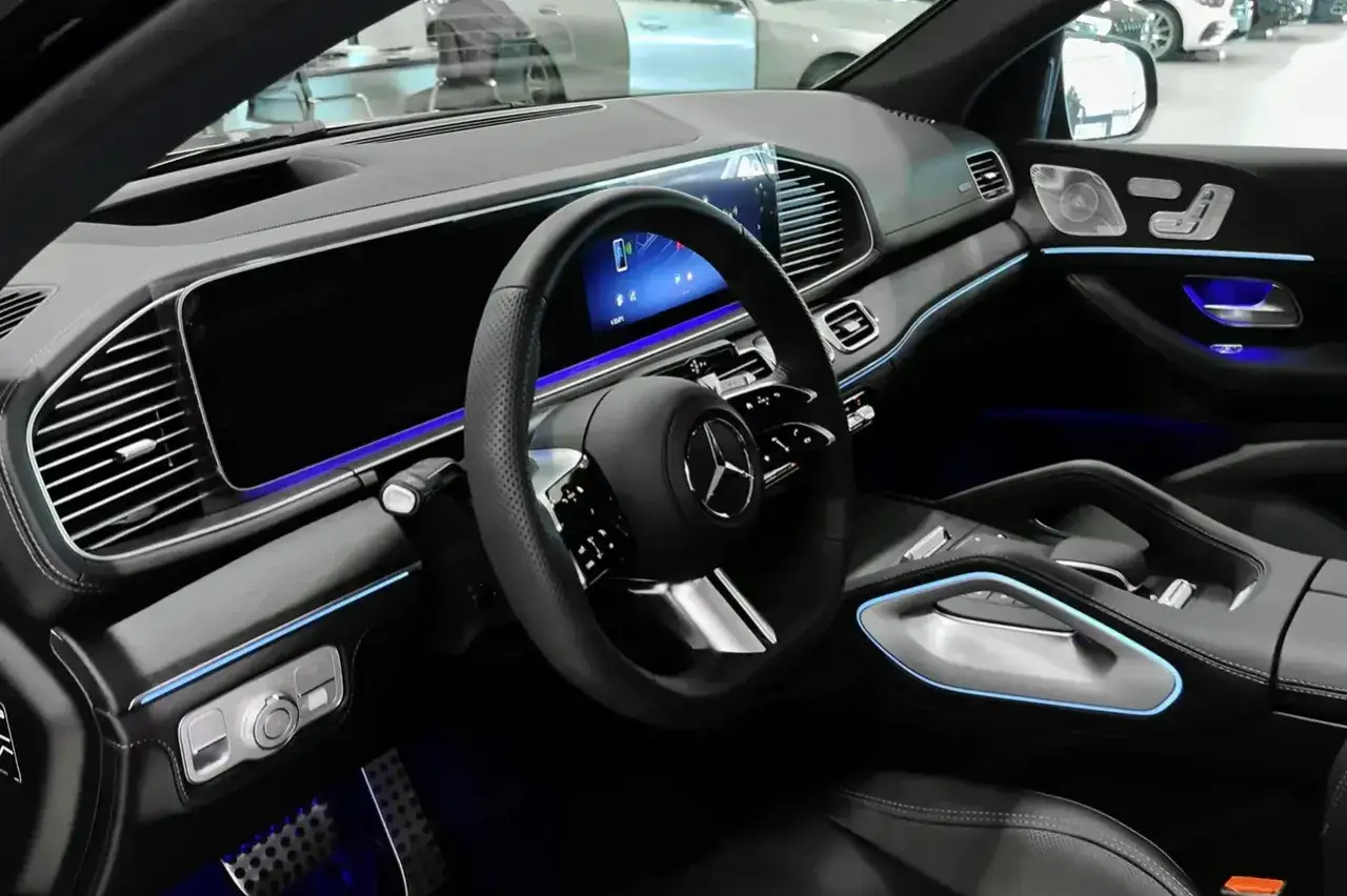 Wnętrze nowoczesnego Mercedesa GLE 2025 z podświetleniem ambientowym. Widoczna kierownica, deska rozdzielcza z ekranami i konsola środkowa.