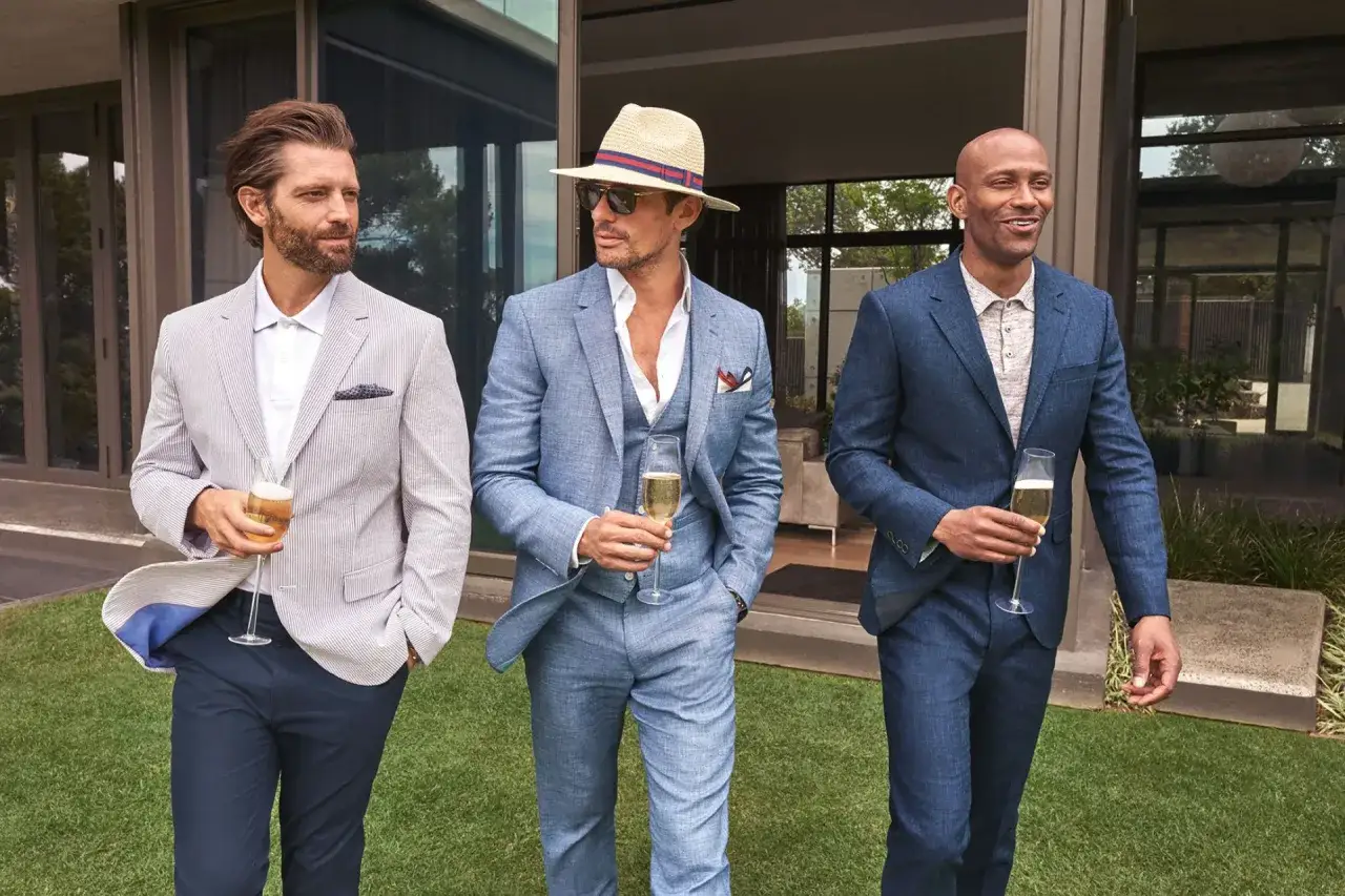 Three men in stylish no suit wedding attire, holding champagne glasses, enjoy a sunny day outdoors.