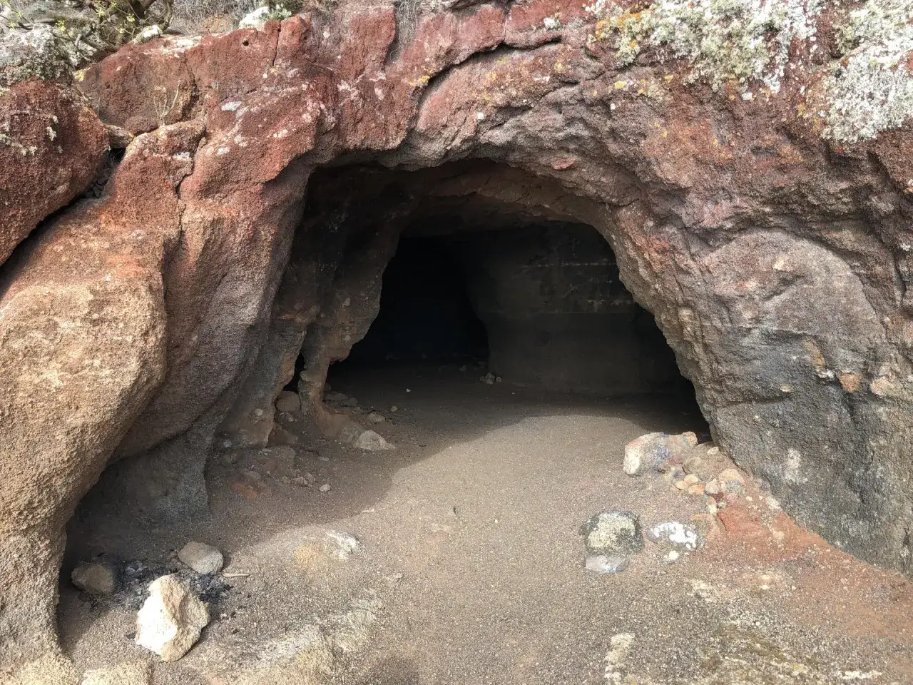Entrada oscura a una cueva bermeja, con rocas rojizas y suelo arenoso.