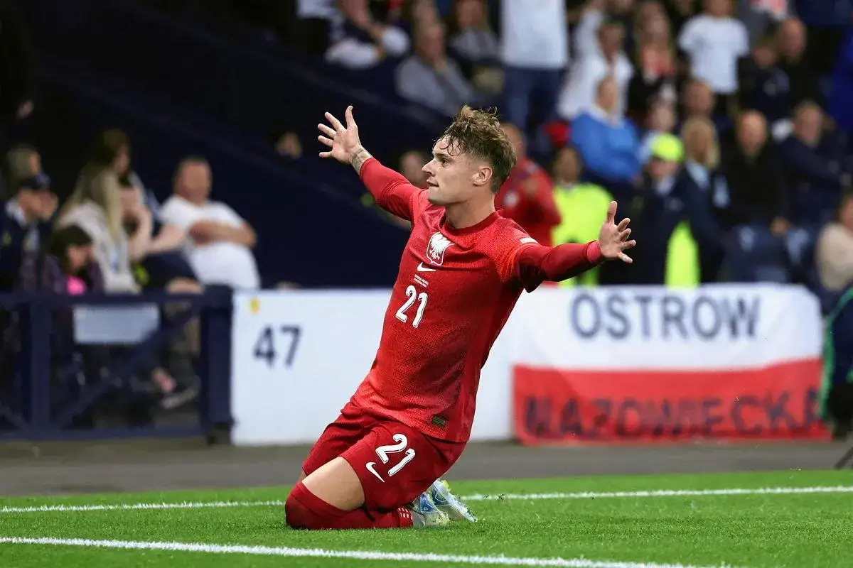 Piłkarz w czerwonej koszulce z numerem 21, Sebastian Szymański, klęczy na boisku z rękami uniesionymi w geście radości. Jego **wzrost** formy jest widoczny w tej dynamicznej pozie.