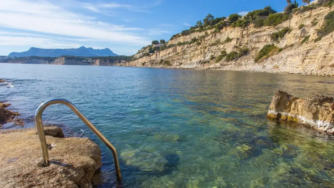 Escalera metálica para acceder a las aguas cristalinas de **Punta Estrella Moraira**, con acantilados y el mar azul de fondo.
