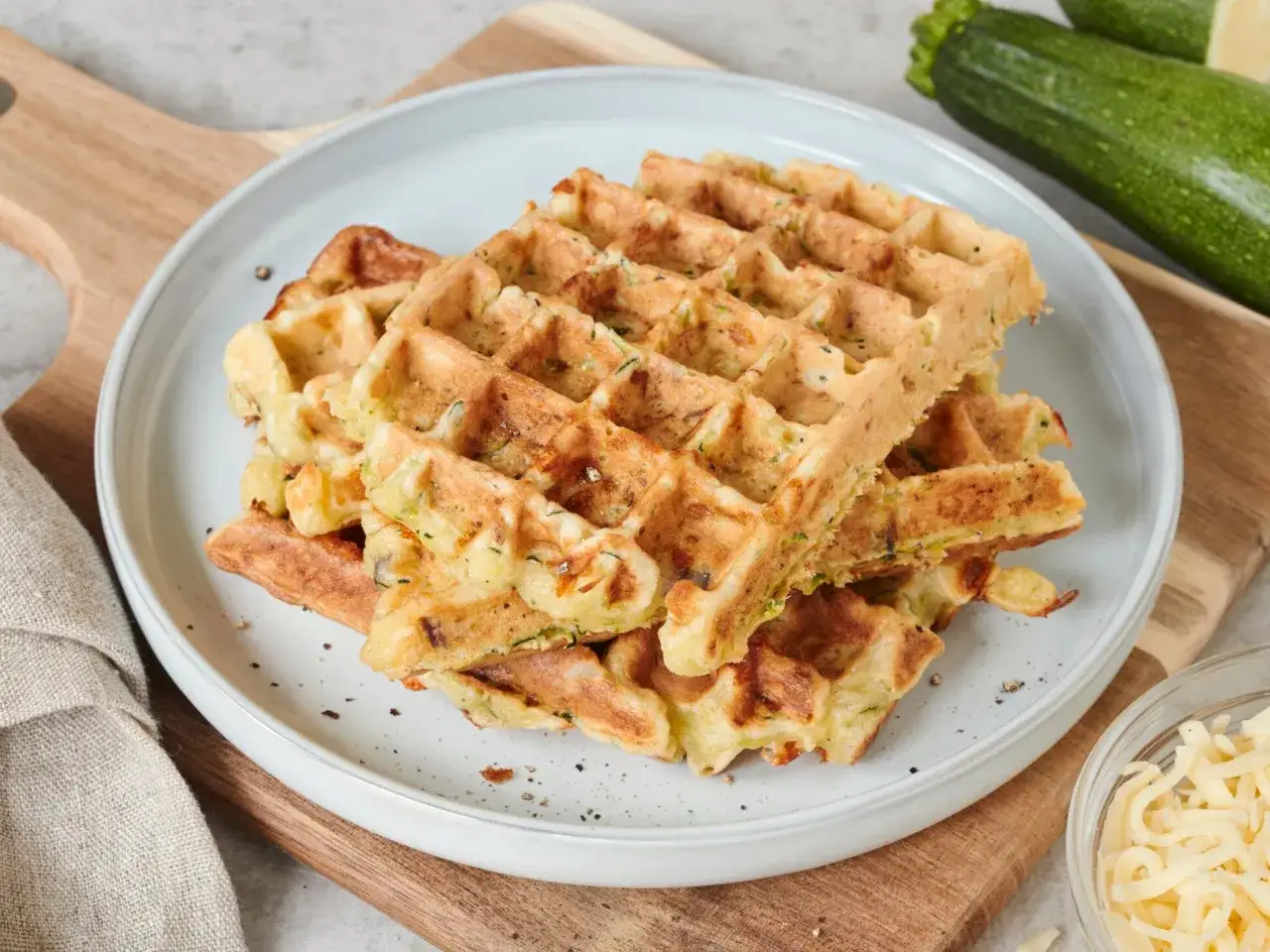 Goldbraune zucchini waffeln auf einem Teller, daneben frische Zucchini und geriebener K&auml;se.