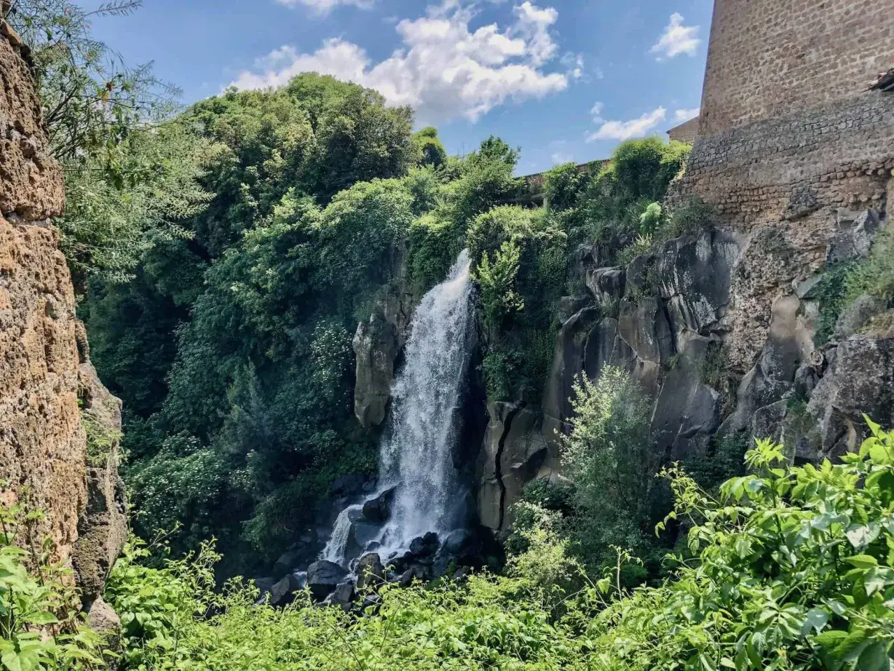 Cascate di Nepi, un'oasi verde con acqua che scende tra rocce e vegetazione rigogliosa, sotto un cielo azzurro con nuvole bianche.