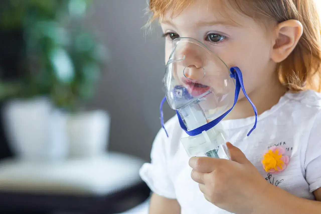 Mała dziewczynka z rudymi włosami korzysta z inhalatora. Jej twarz jest częściowo zakryta przez maskę inhalacyjną, przez którą wdycha sól fizjologiczną.
