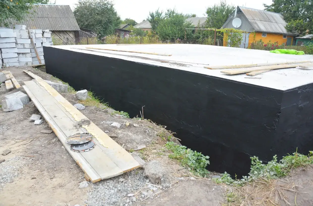 Schwarze Abdichtung der Bodenplatte schützt vor hohem Grundwasserspiegel. Baustelle mit Betonsteinen und Holzbrettern.