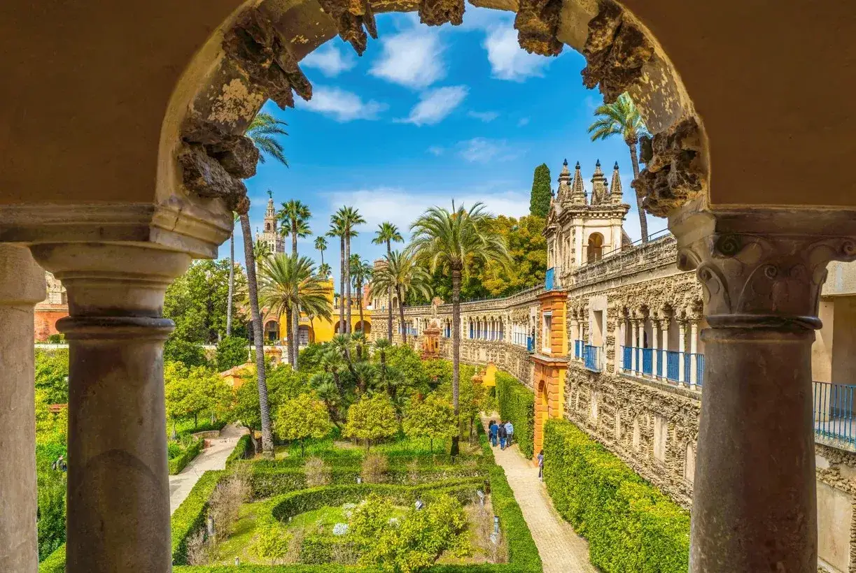 Blick durch einen Torbogen auf die G&auml;rten des Alc&aacute;zar Sevilla mit Palmen, Orangenb&auml;umen und einer pr&auml;chtigen Architektur.