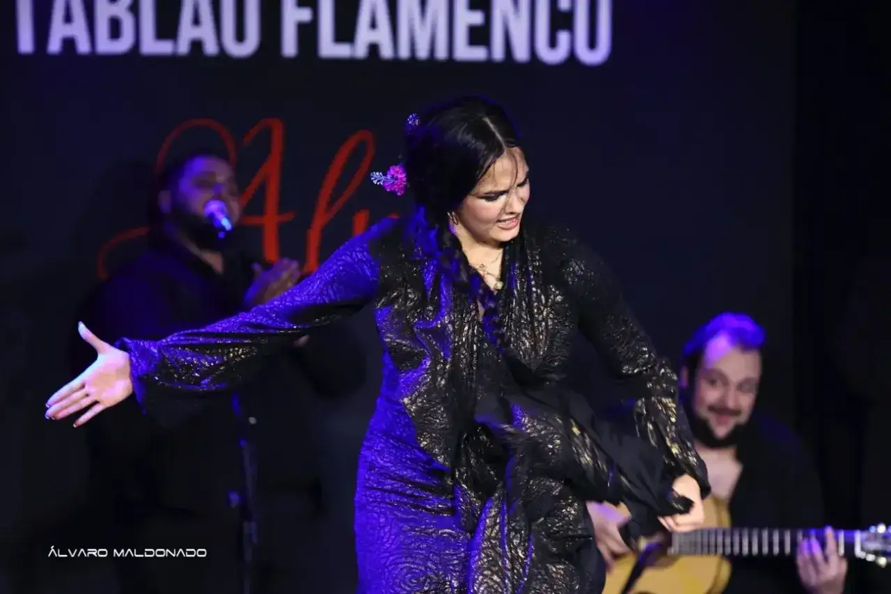 Bailarina de flamenco con vestido negro brillante, moviendo sus brazos al ritmo de la rumba española. Un guitarrista la acompaña.
