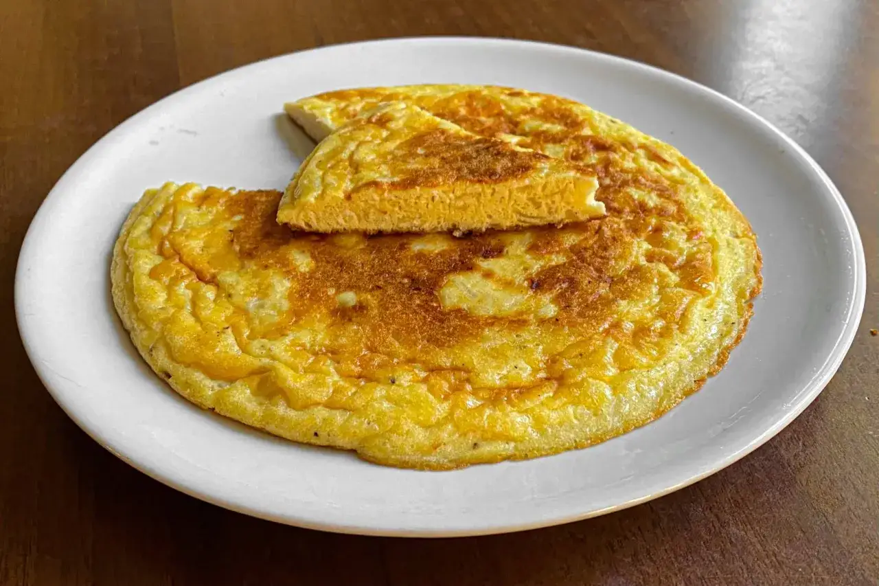Frittata di cipolle dorata e soffice, servita su un piatto bianco. Una fetta &egrave; stata sollevata, mostrando la consistenza interna.