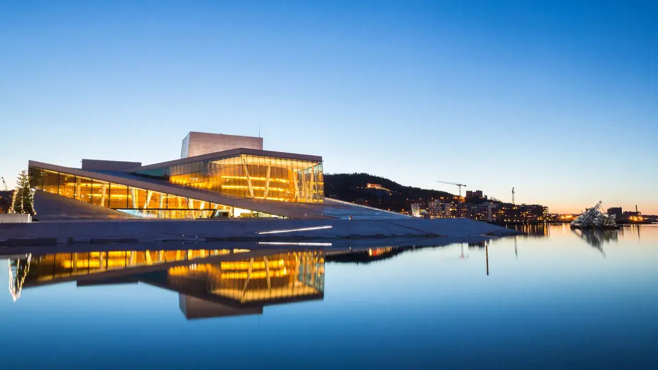 Oslo w jeden dzień: nowoczesna Opera odbija się w spokojnej wodzie o zmierzchu. W tle widać oświetlone miasto i wzgórza.