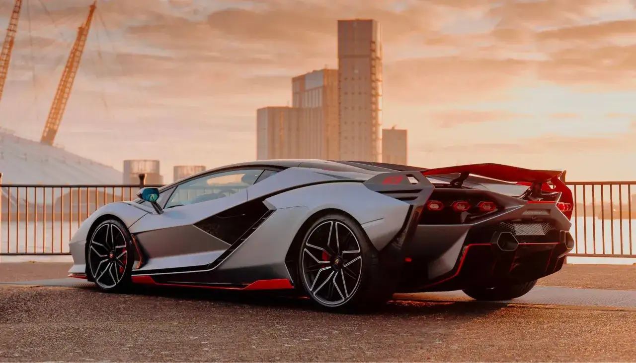 Srebrny Lamborghini Sian z czerwonymi akcentami na tle zachodzącego słońca i nowoczesnej architektury.