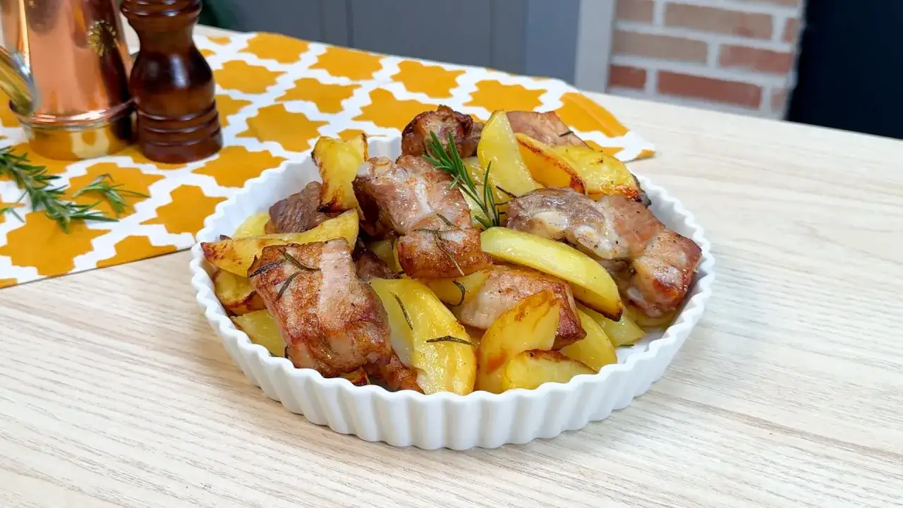 Succulente costine al forno con patate dorate e rosmarino, servite in una ciotola bianca su un tavolo di legno.
