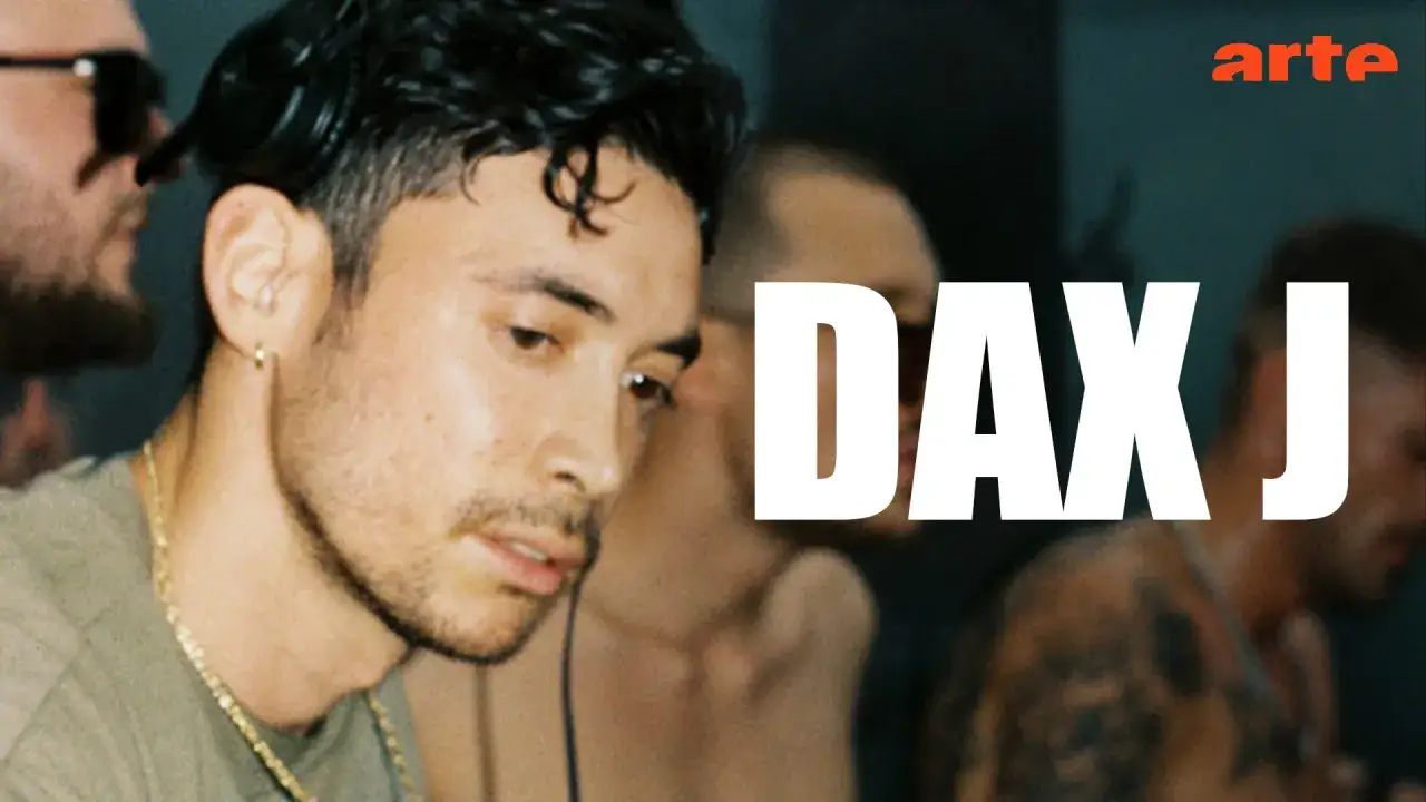 Dax J, artysta z charakterystycznym kolczykiem w uchu i złotym łańcuchem, w otoczeniu innych os&oacute;b.