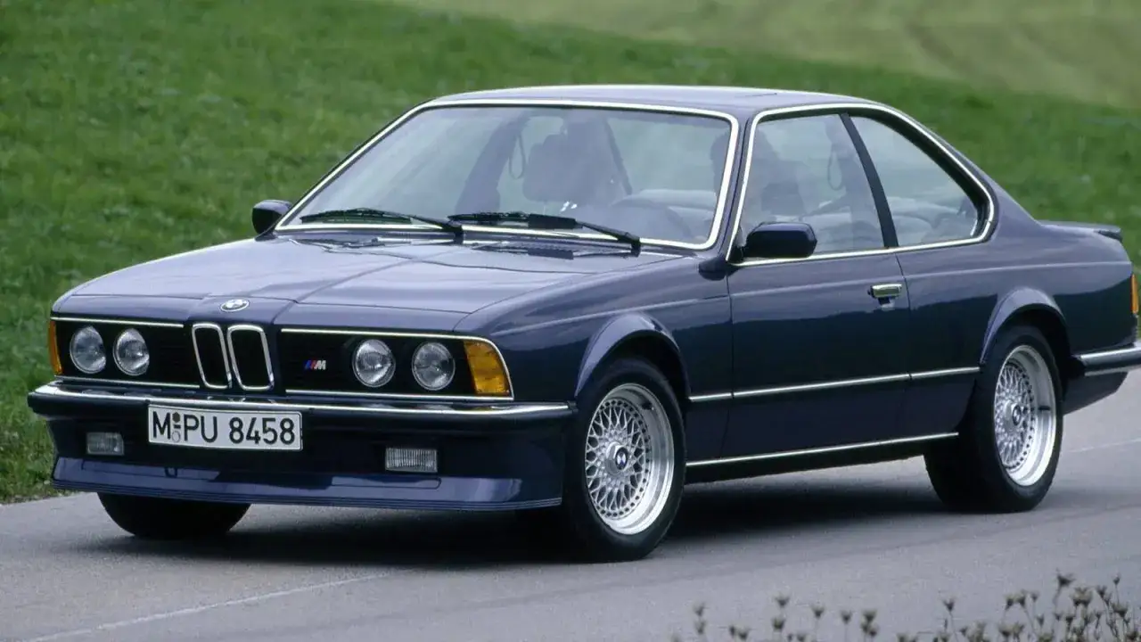 M&ouml;rkbl&aring; BMW 80-tals coup&eacute; med kromdetaljer och klassiska f&auml;lgar, parkerad p&aring; en asfalterad v&auml;g med gr&ouml;n bakgrund.