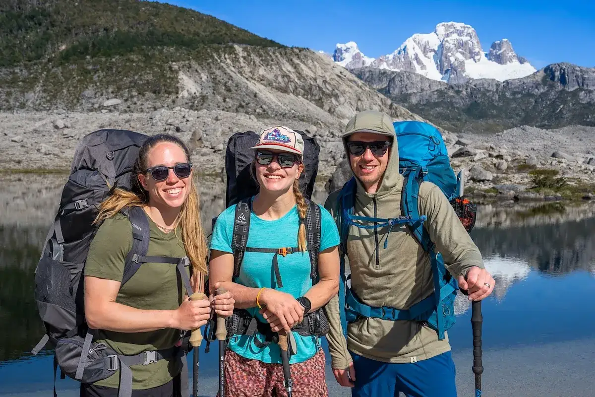 Tres excursionistas sonríen con mochilas y bastones, listos para la aventura. La mujer de la izquierda viste una camiseta de **merino material**.