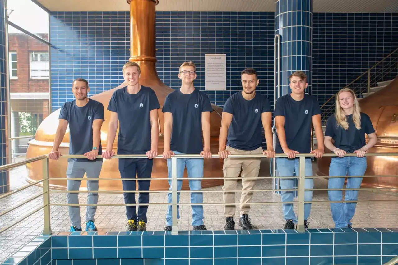 Sechs junge Leute in dunklen T-Shirts stehen vor gro&szlig;en Kupferkesseln. Sie machen ihre Ausbildung zum Brauer und sind stolz auf ihren Beruf.