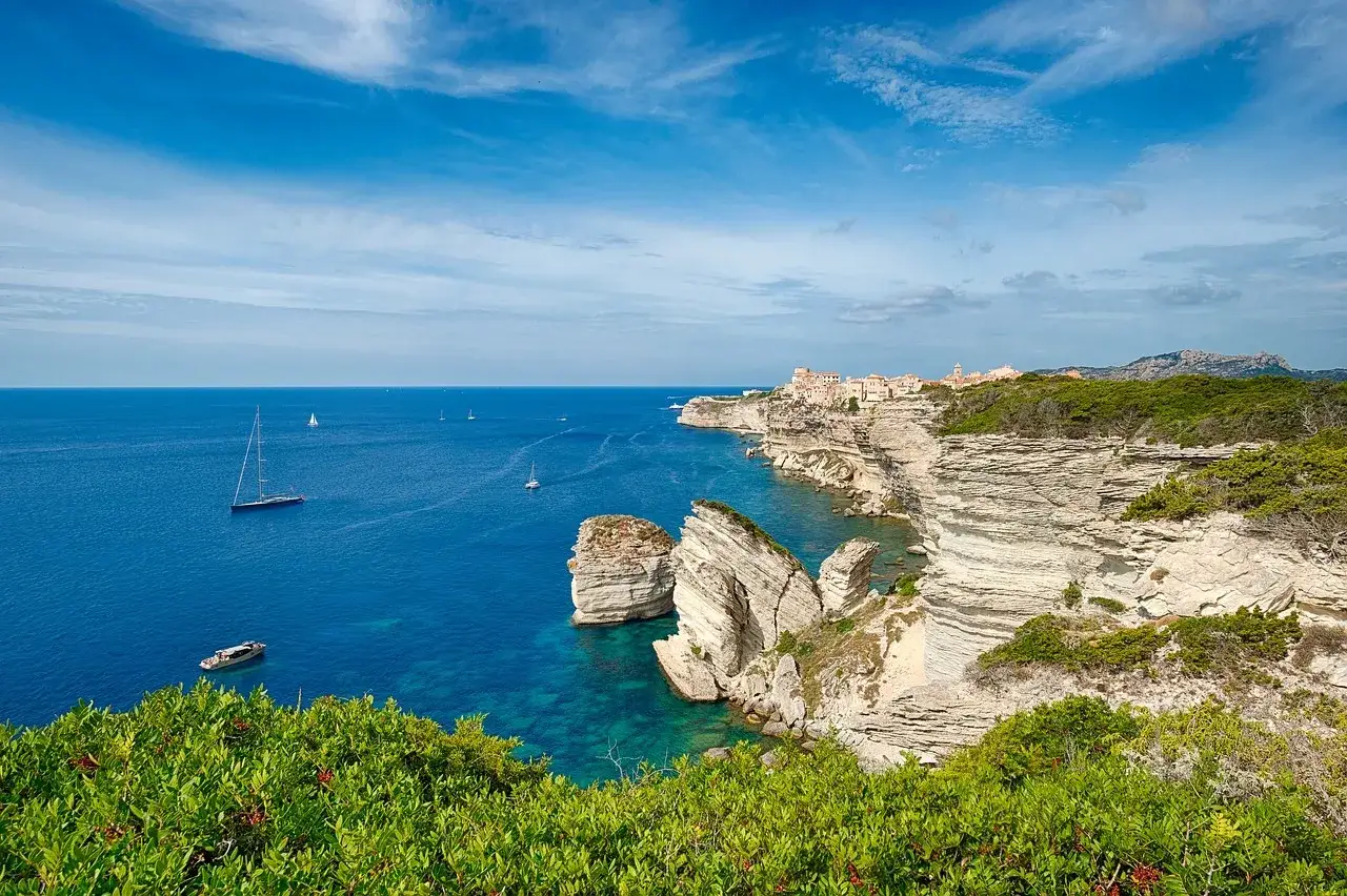 Blick auf Bonifacio, Korsika: Weiße Felsklippen ragen steil aus dem türkisfarbenen Meer auf, darauf eine Stadt. Segelboote kreuzen auf dem blauen Wasser.