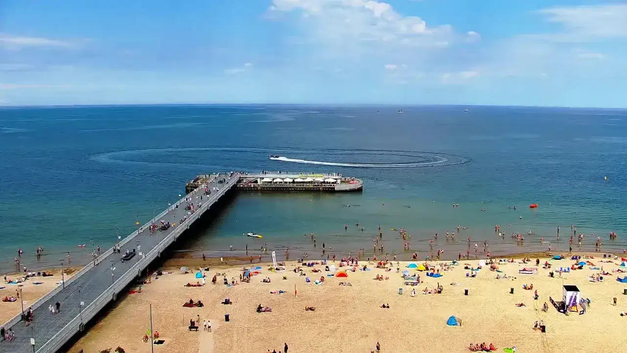 Słoneczna kołobrzeg plaża tętni życiem. Ludzie spacerują po molo, kąpią się w morzu i odpoczywają na piasku.