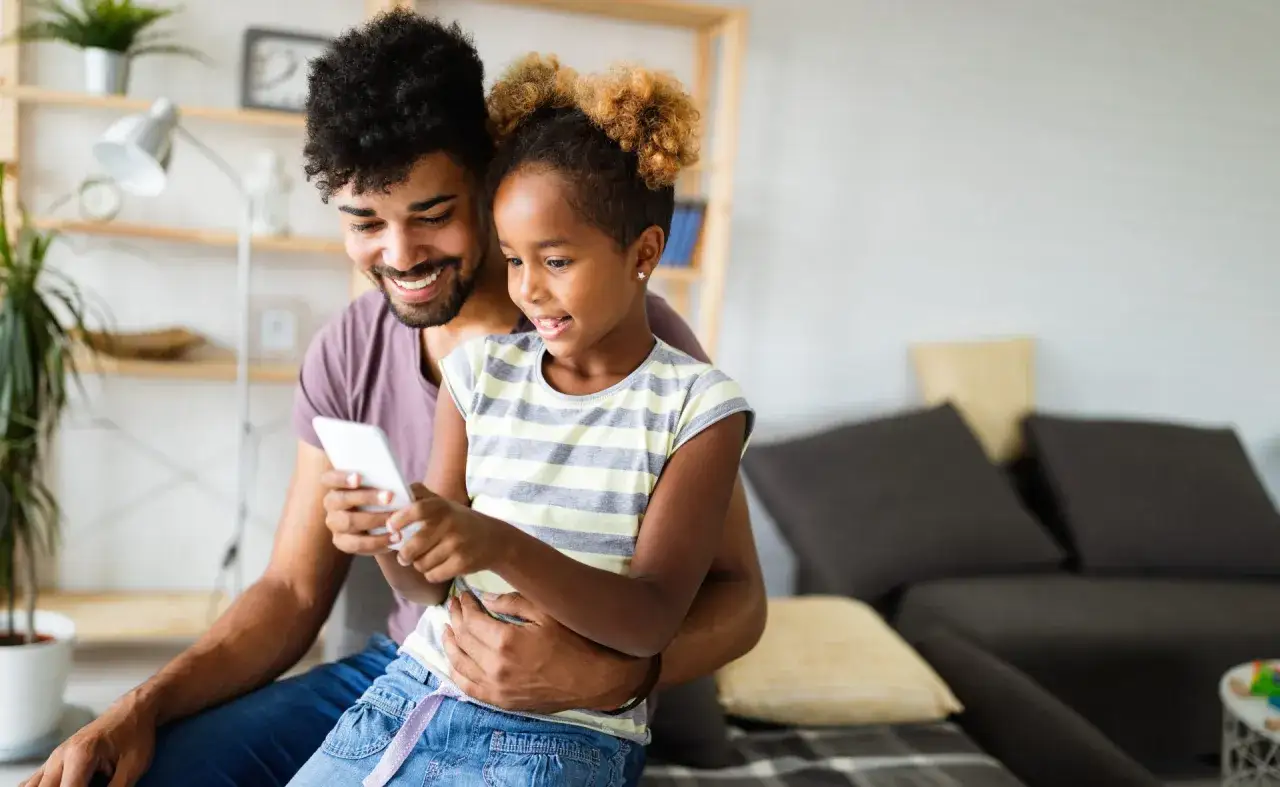 Padre e hija sonríen mirando un móvil, quizás explorando la cuenta infantil BBVA para aprender sobre finanzas de forma divertida.