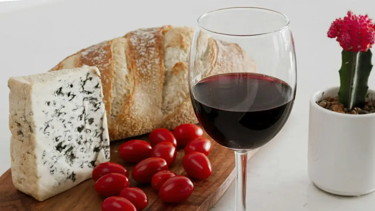 A glass of Alicante Bouschet wine sits beside a wedge of blue cheese, crusty bread, and cherry tomatoes on a wooden board.