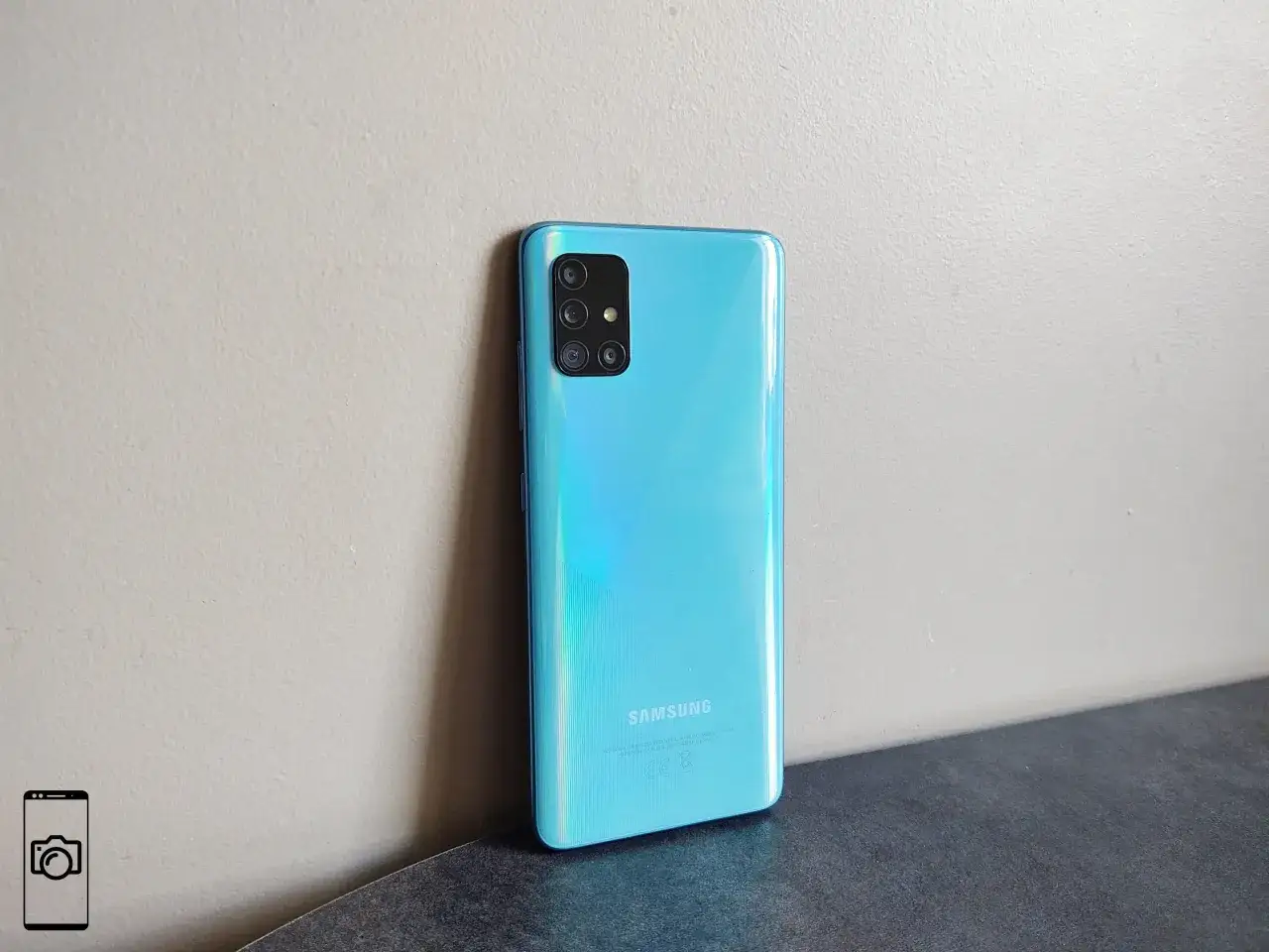 Błękitny Samsung A51 z aparatem z czterema obiektywami oparty o jasną ścianę.