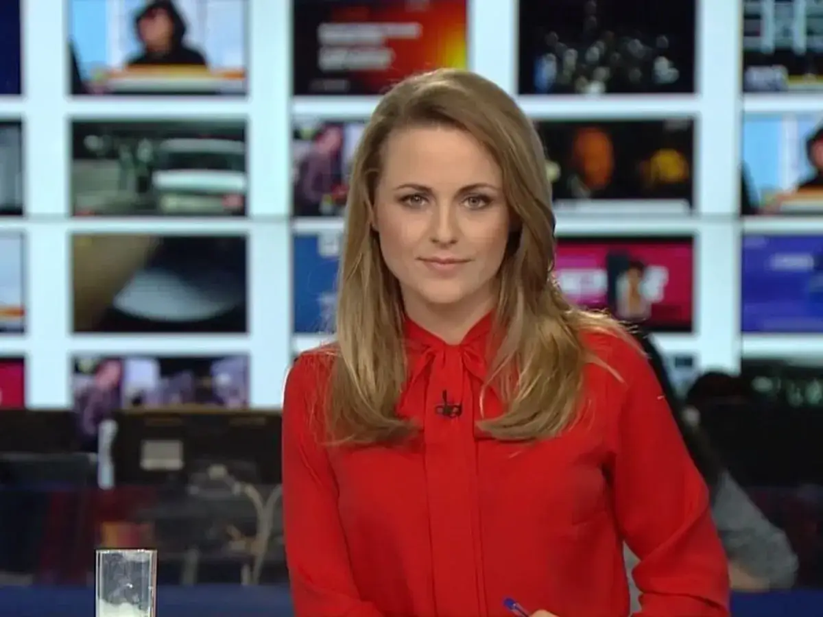 Marta Kłos w czerwonej bluzce przed ekranami TVN Fakty.
