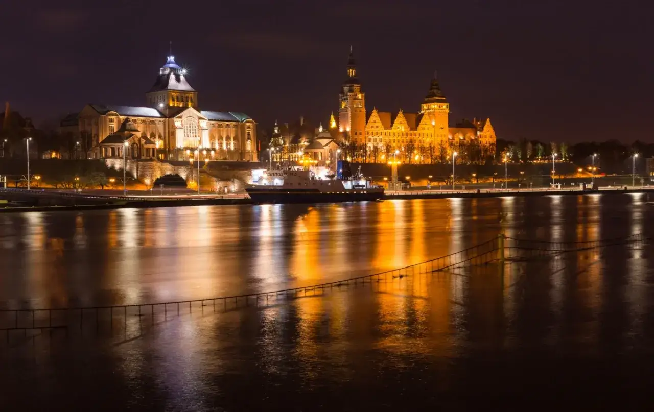 Nocny widok na oświetlone budynki i rzekę w Szczecinie. Statek kołysze się na wodzie, a odbicia świateł tworzą magiczny klimat, idealny na nietypowe atrakcje Szczecin.