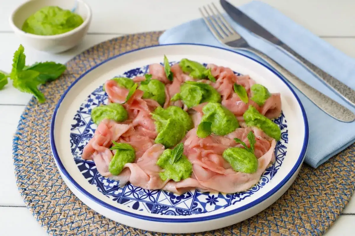 Fettine rosa di carpaccio con quenelle di crema verde e foglie di menta, un piatto che ricorda le zucchine alla scapece per freschezza.