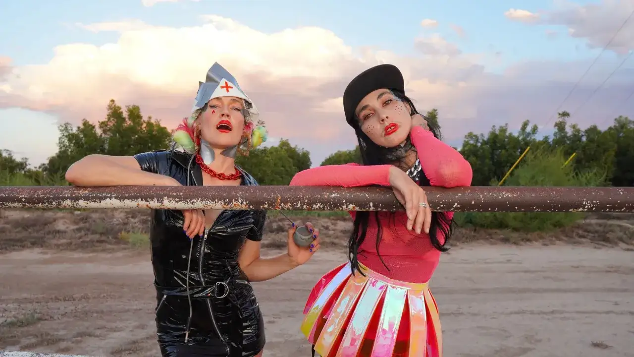 Deux femmes au look audacieux, inspiré de coco rosie, posent devant un ciel pastel. L'une porte une tenue en vinyle noir, l'autre une jupe irisée.