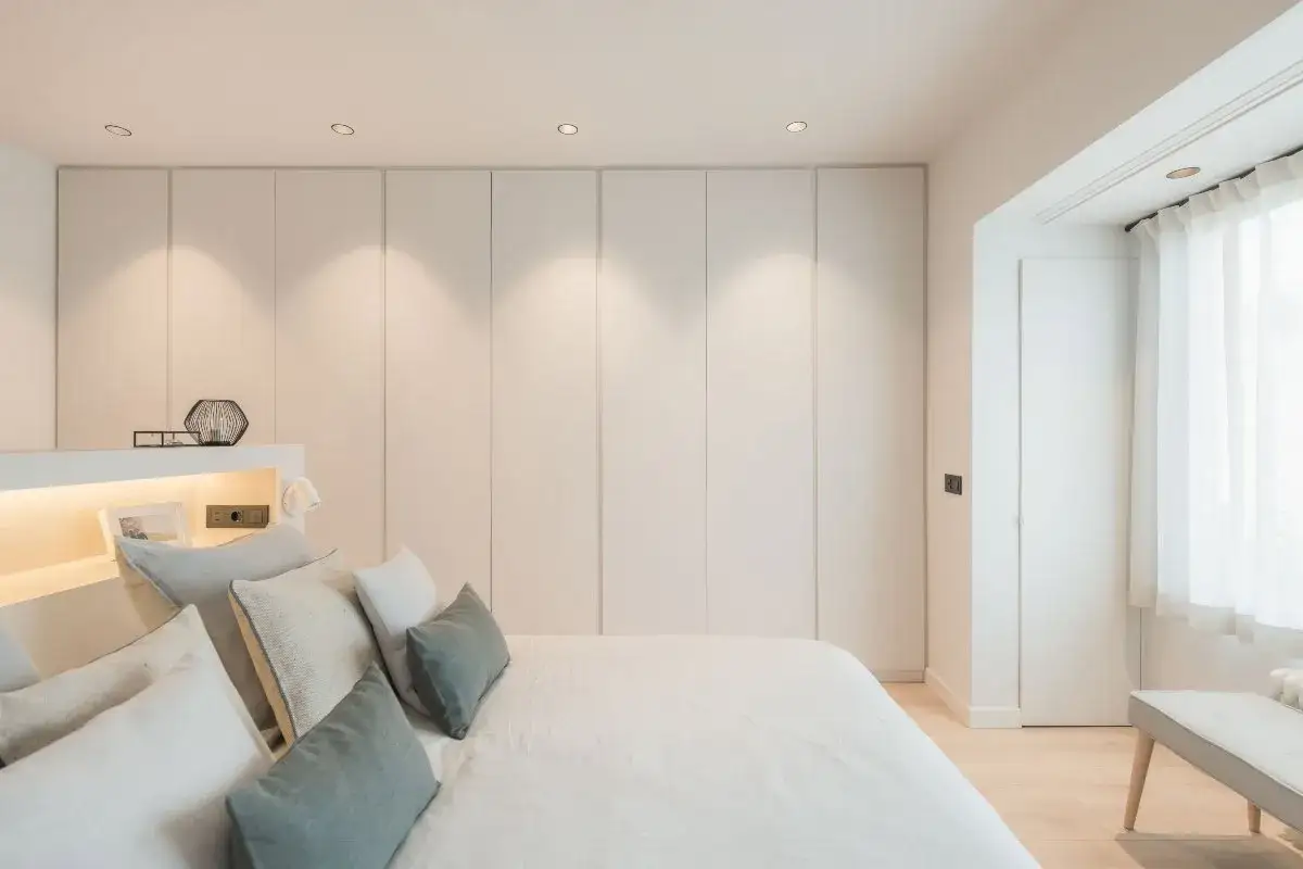 Dormitorio minimalista con cama y almohadas. Armarios blancos integrados aprovechan hueco entre cama y pared. Luz tenue ilumina la escena.