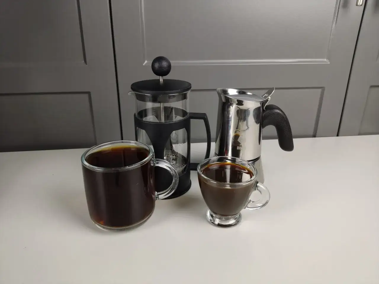 Två koppar kaffe, en presskanna och en mokabryggare står på ett vitt bord. Man kan brygga presskaffe i kaffebryggare, men här ser vi olika sätt att njuta av kaffe.