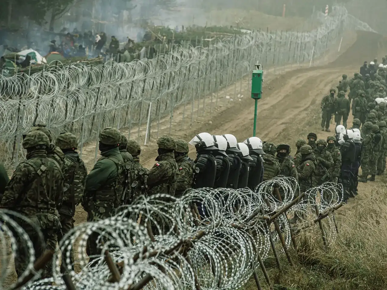 Żołnierze i policjanci stoją za zasiekami z drutu kolczastego, pilnując granicy. W tle widać obóz migrantów, symbolizujący trudną sytuację związaną z imigracją.