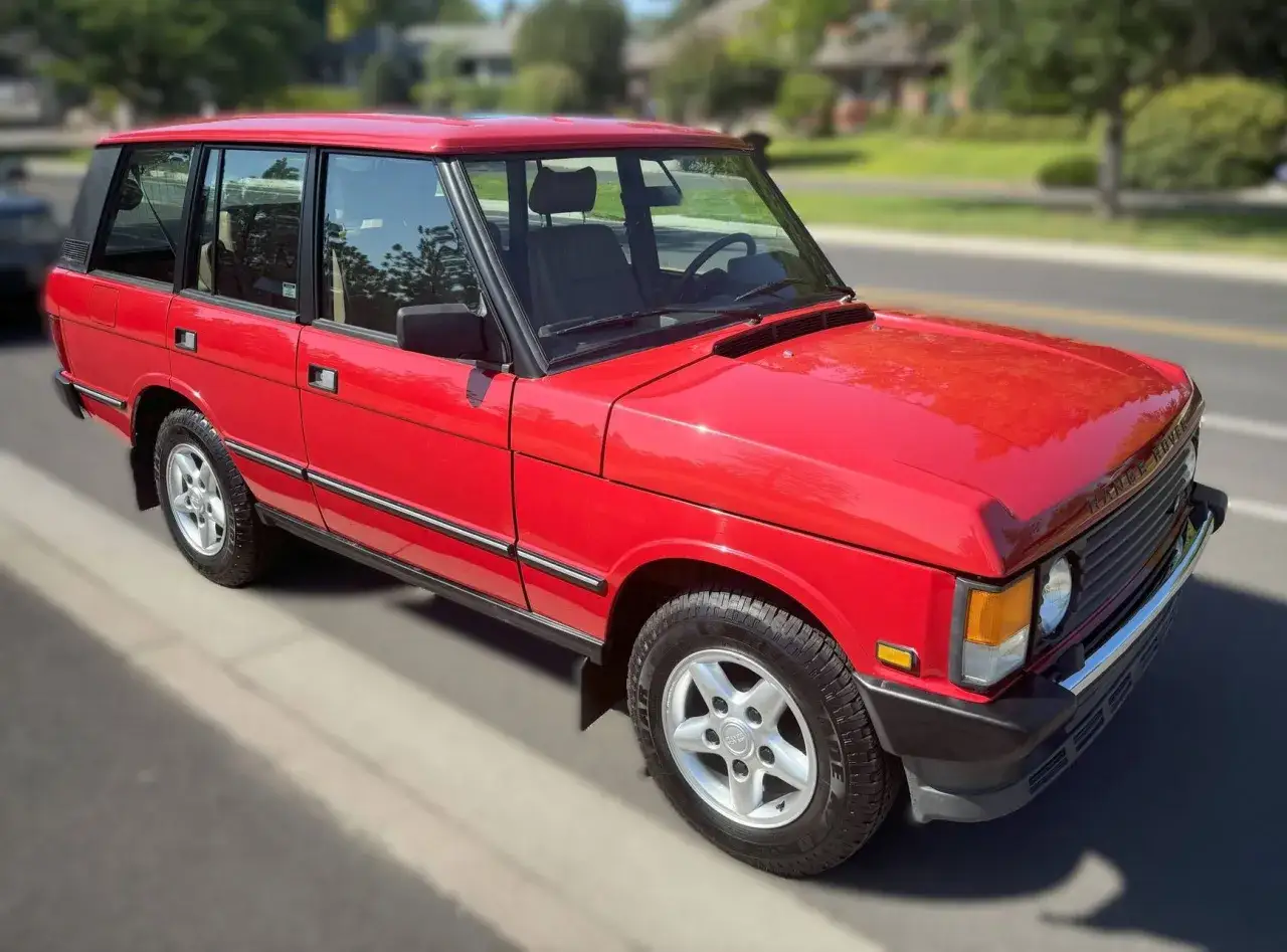 Ein klassischer, knallroter Range Rover steht am Stra&szlig;enrand. Sein gl&auml;nzender Lack reflektiert die Sonne, w&auml;hrend die markanten Linien seine zeitlose Eleganz unterstreichen.