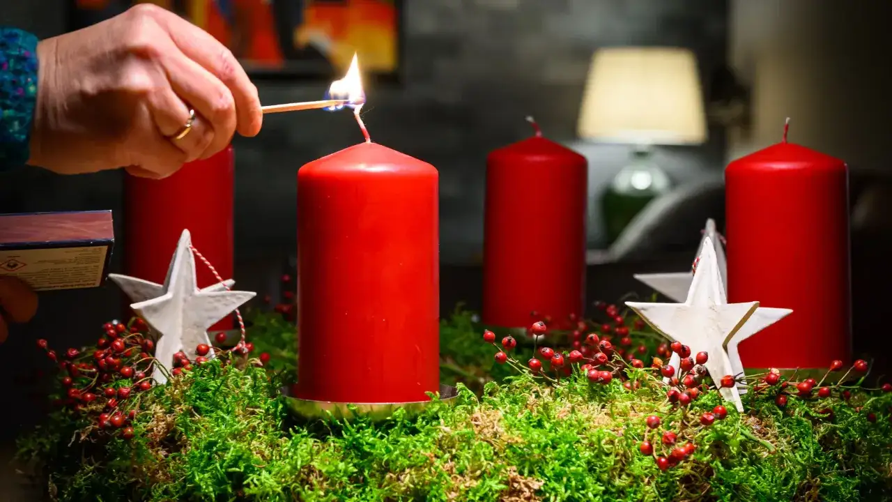 Eine Hand zündet die erste Kerze eines Adventskranzes an. Rote Kerzen, Moos und Beeren schmücken den Kranz, ein Symbol deutscher Weihnachtstraditionen.