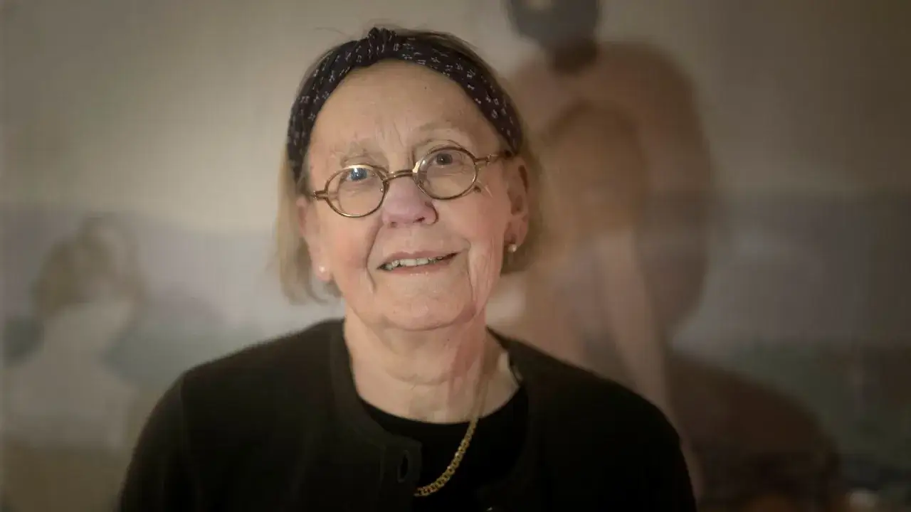 Anna Asp, scenograf, med glasögon och pannband, ler mot kameran. Bakgrunden är suddig med en figur.