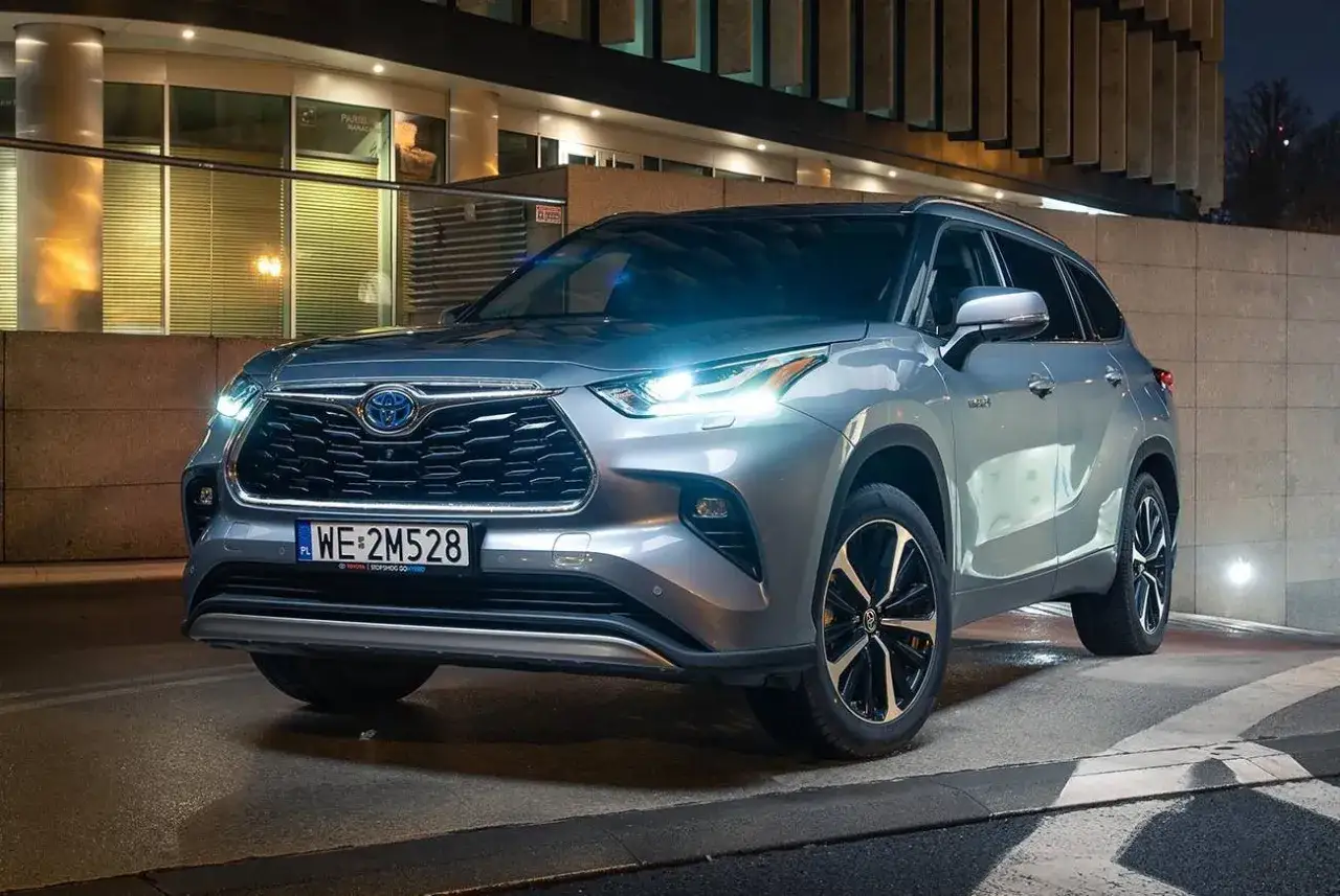 Srebrny Toyota Kluger z włączonymi światłami, zaparkowany nocą przed nowoczesnym budynkiem.