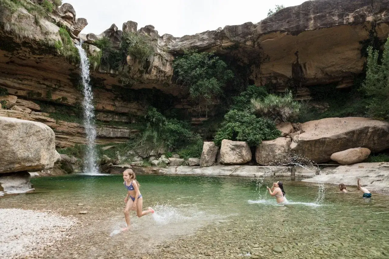 Ni&ntilde;os disfrutan en unas **piscinas naturales** bajo una cascada, rodeados de rocas y vegetaci&oacute;n.