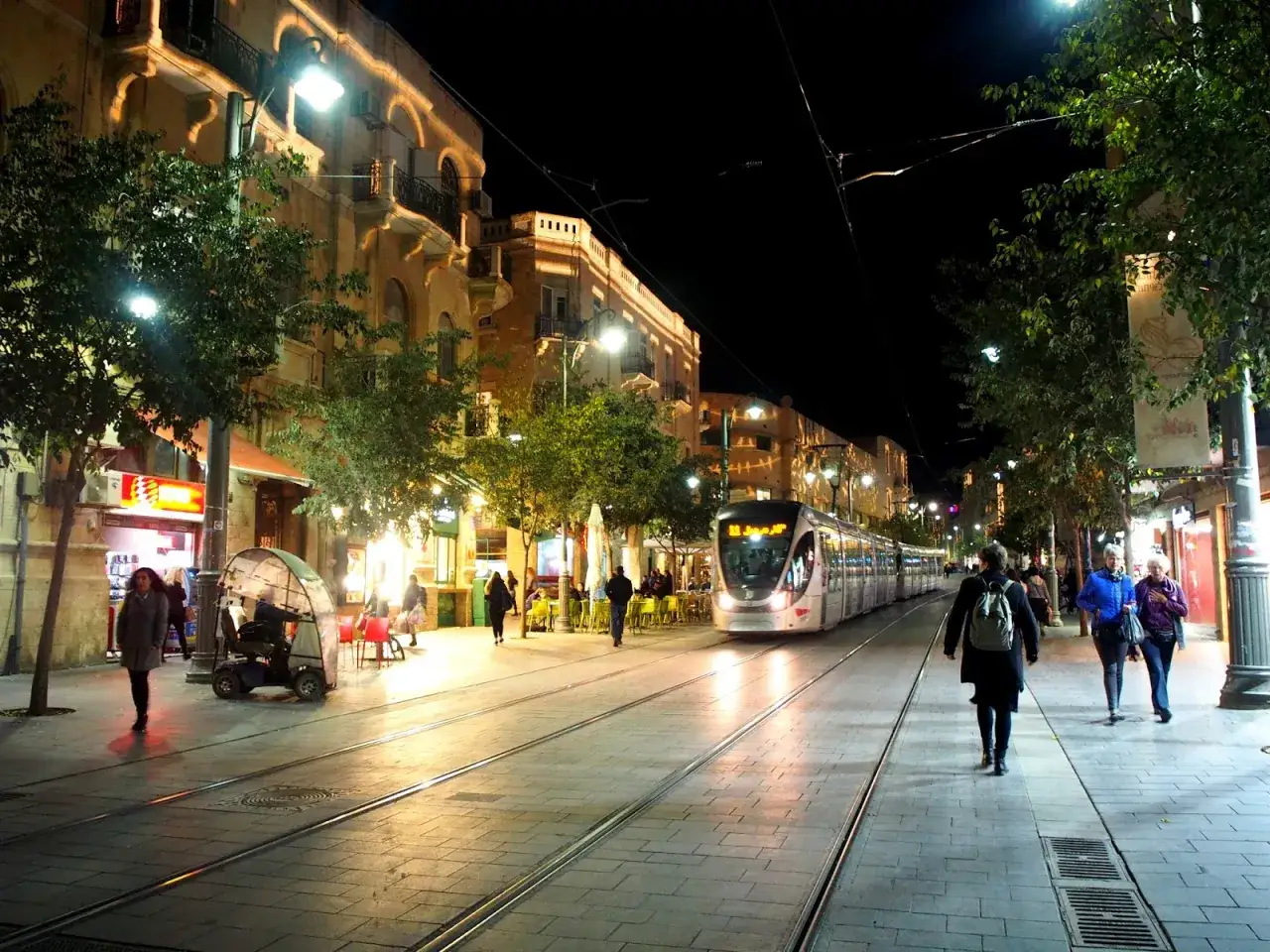 Nocne życie w jednym z izrael miasta. Tramwaj przejeżdża obok oświetlonych budynków i drzew, a ludzie spacerują po chodniku.
